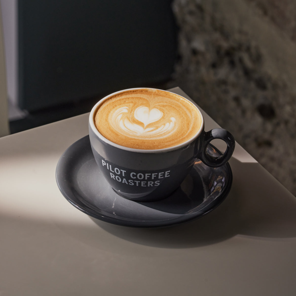 Close up shot of a latte with heart art in a Pilot Coffee Roasters branded cup and saucer on a grey worktop. 