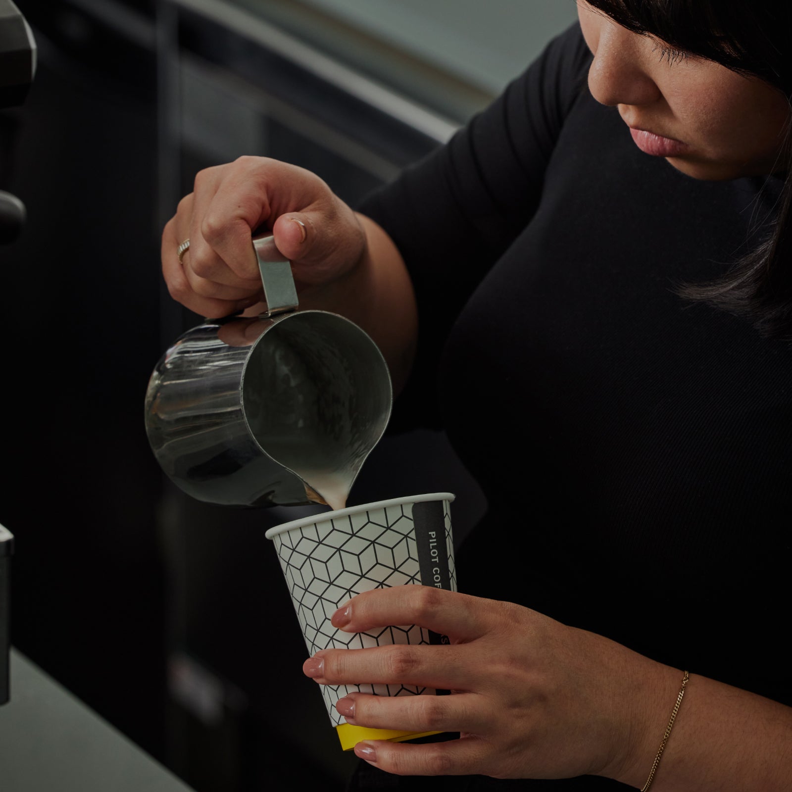 A barista pouring milk into a Pilot Coffee Roasters branded takeaway cup. The angle of the cup suggests she is creating latte art