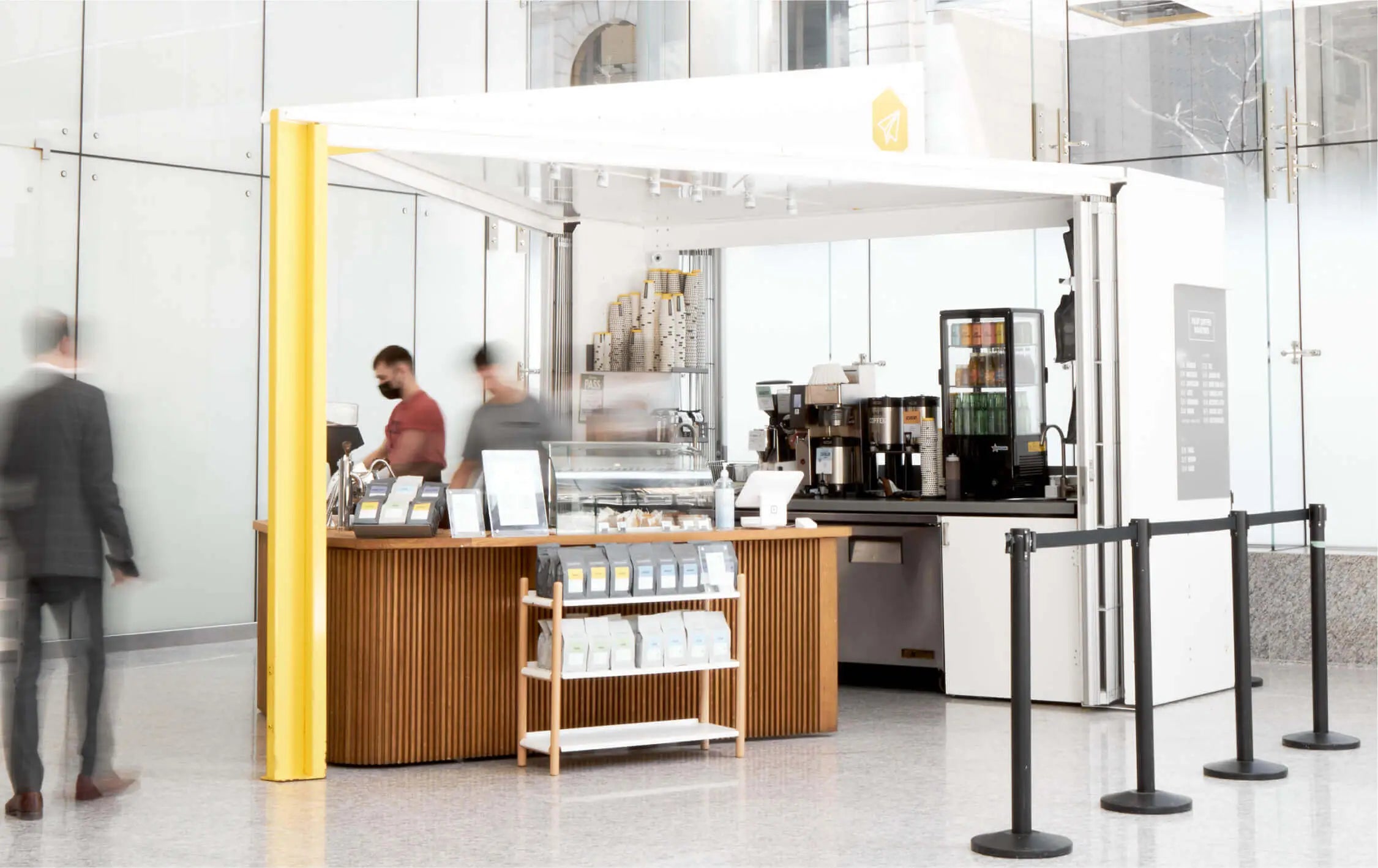 A brightly lit shot of the Bay Adelaide Center Kiosk. Two Barista prep drinks behind the bar while a blurred customer moves towards the kiosk. The wall walls of the building stream in light in the background