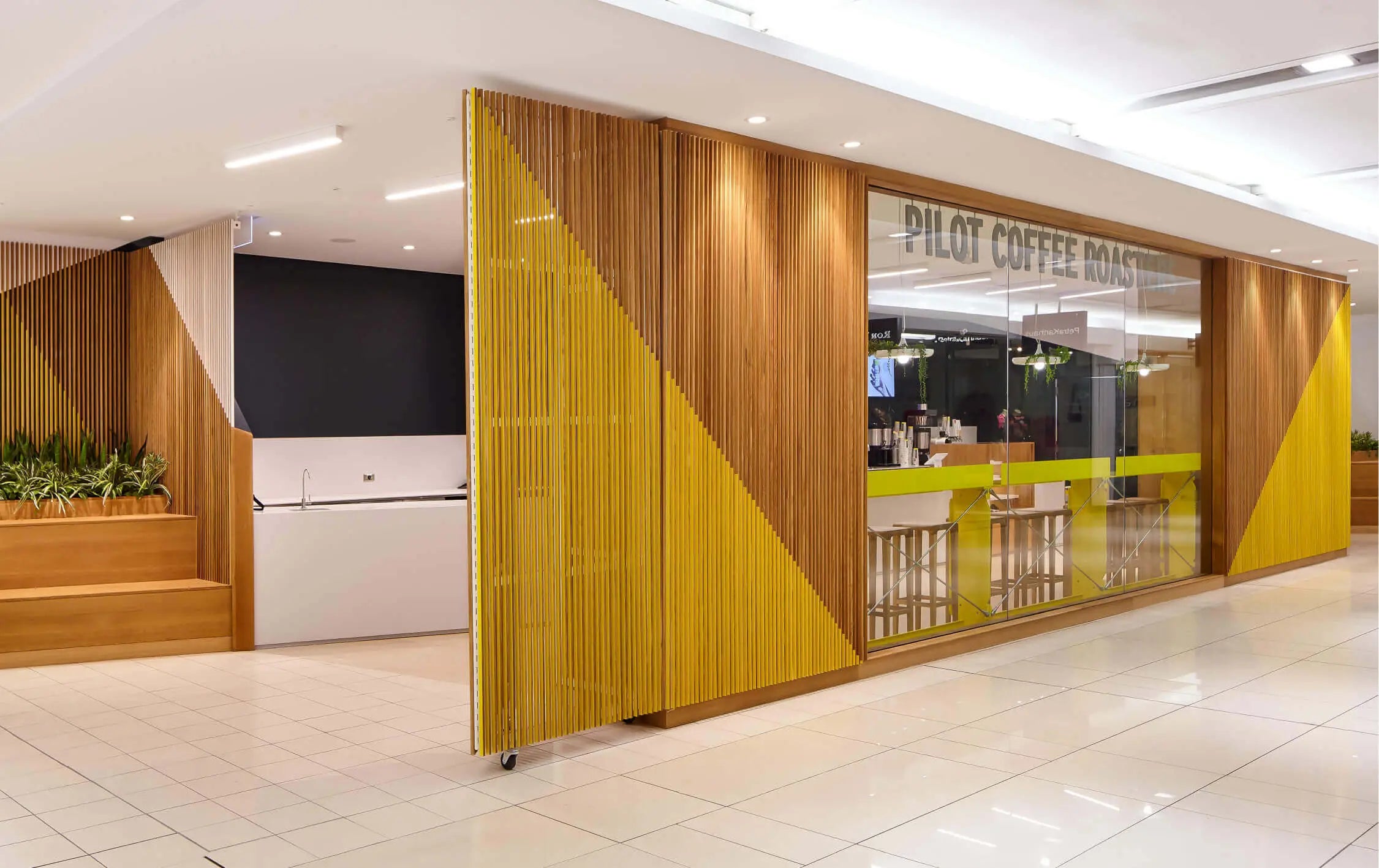 The front of the Bay Bloor Cafe in the Manulife Center The yellow and natural wood slats surround a glass window with the Pilot Coffee Roasters word mark on top. The cafe is visible through the glass. Wood bleacher seating can be seen on the left as well as just in frame on the right.