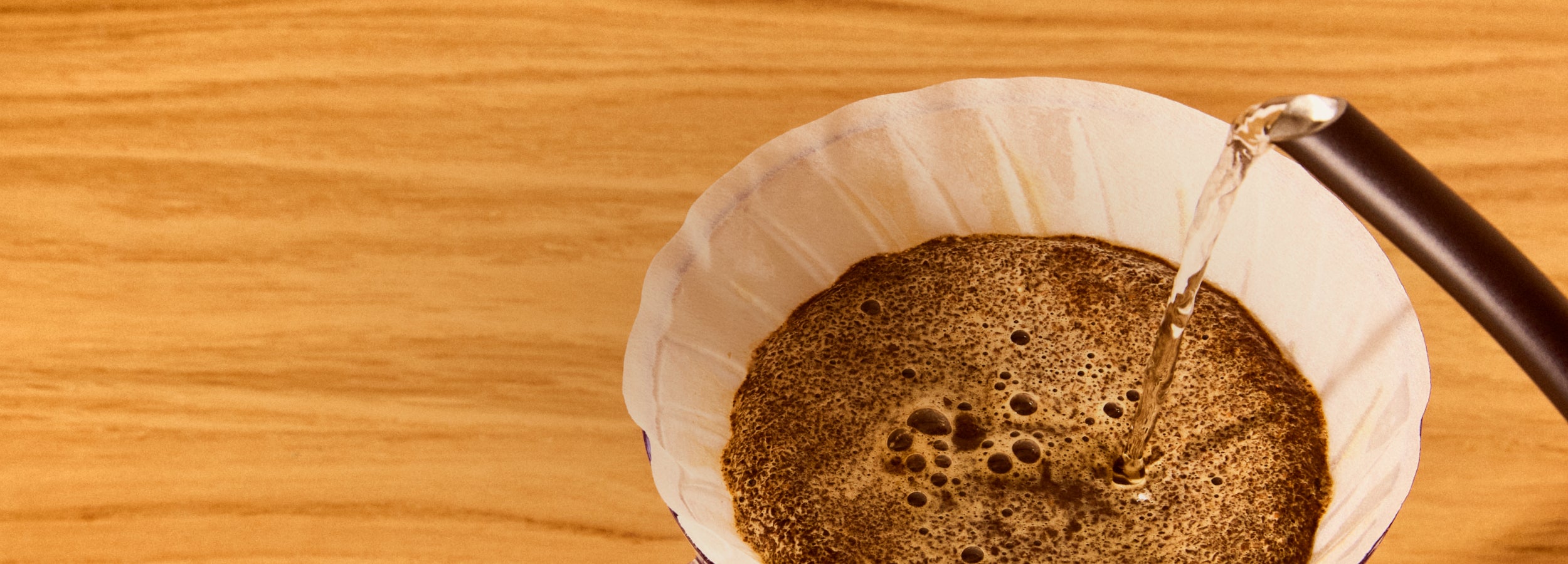 Close up shot of a gooseneck kettle pouring water over a v60 dripper in it's first pour after bloom on a wooden surface