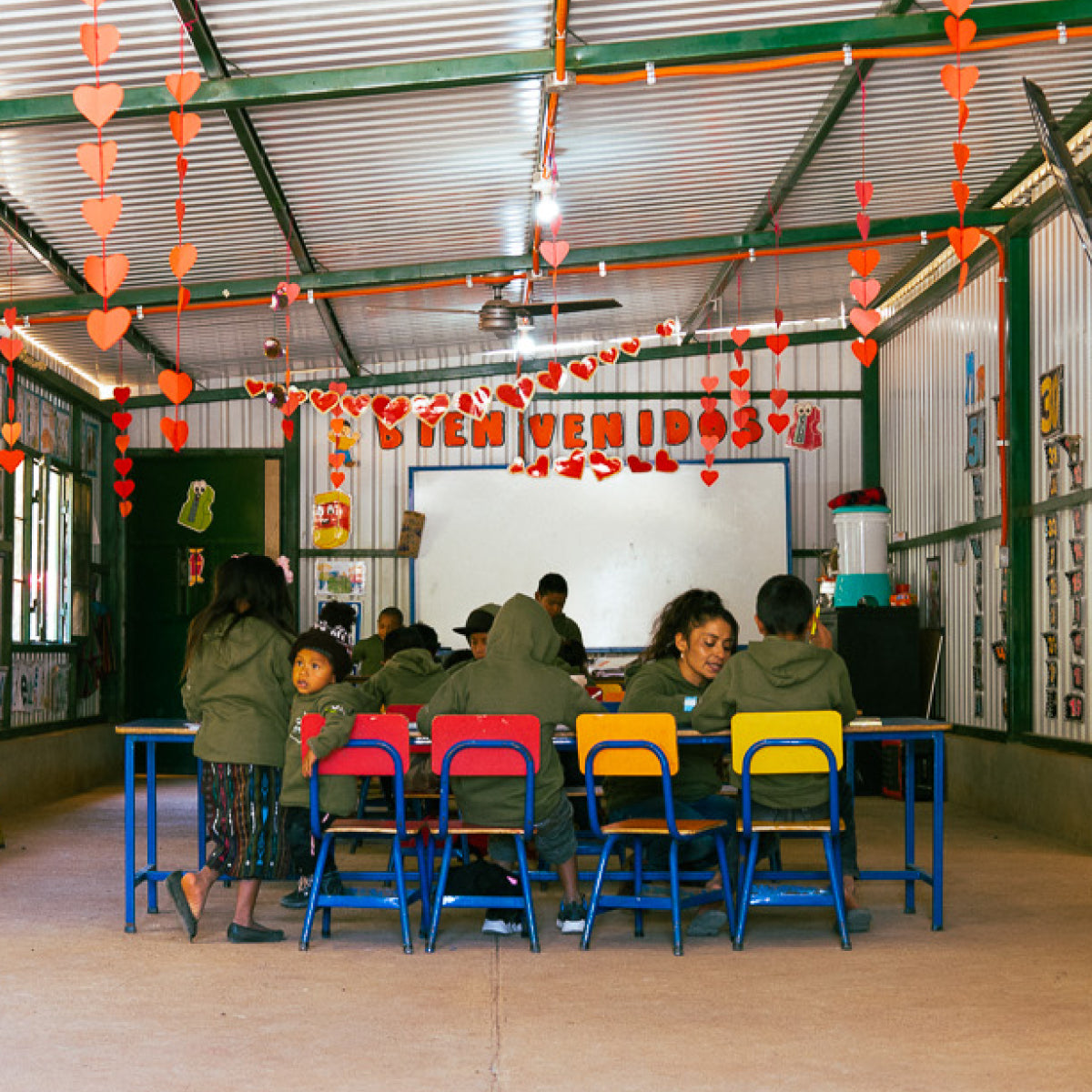 School children and educators sitting on brightly coloured red, orange and yellow chairs and standing around a long work table in the Catalan Education Center. Paper crafts hang from the ceiling and on the walls. A welcome sign that says "Bienvenidos" hangs above the whiteboard in the background