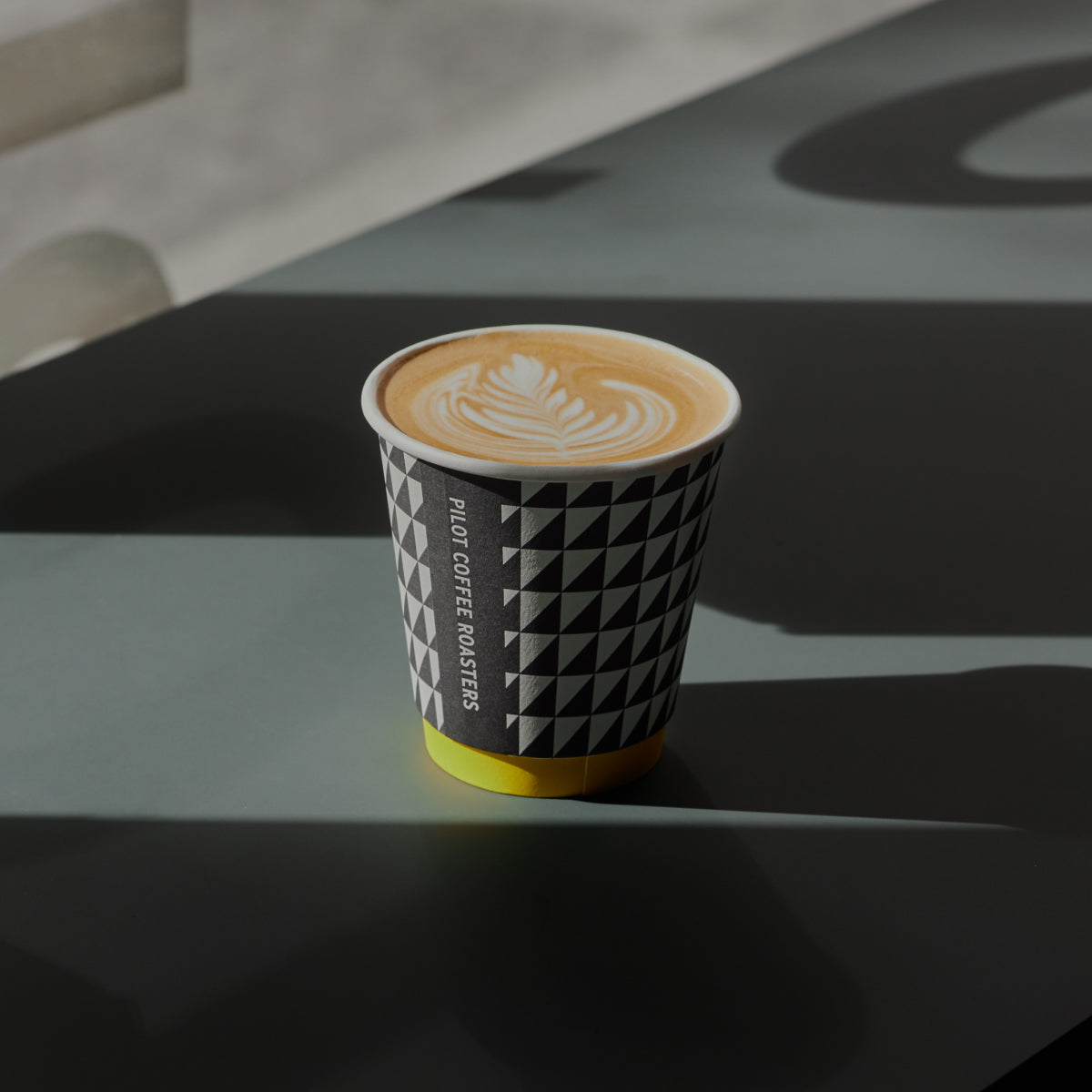 A shadowy image of a Pilot Coffee Coffee Roasters takeway cup on an earthy green countertop. The shadows of letters of the cafe window decals appear on the counter in the background