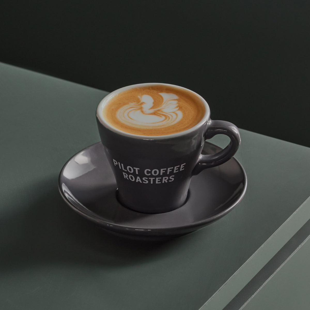 A close up shot of a macchiato with milk art on top in a small Pilot Coffee Roasters branded ceramic cup and saucer. The drink sits atop a green surface with a dark grey wall in the background