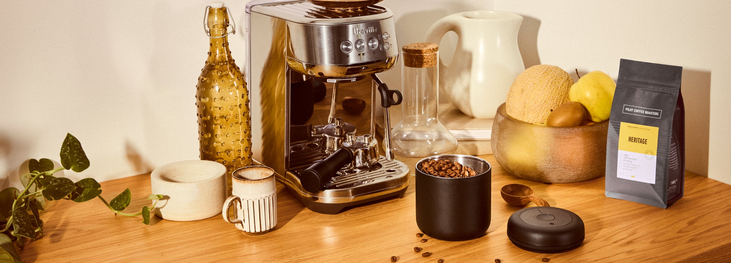 Stylized image of coffee equipment on a wood countertop surrounded by fruits and vases. Including: a Breville Bambino, Fellow Atmos canister, Heritage Blend Coffee and ceramic coffee mugs