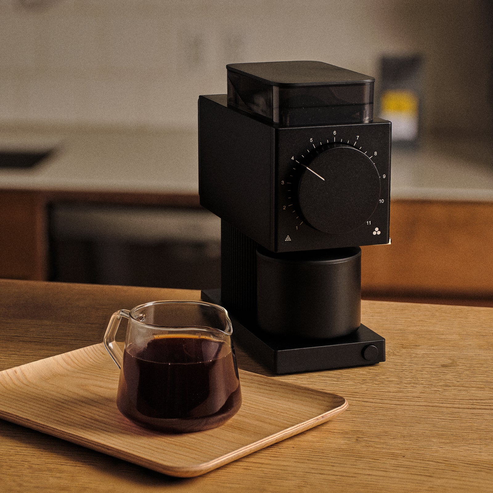 A Fellow Ode Coffee grinder sits on a wood countertop beside a full glass Kinto Carafe. A blurred kitchen worktop and bag of Heritage blend out of focus in the background.