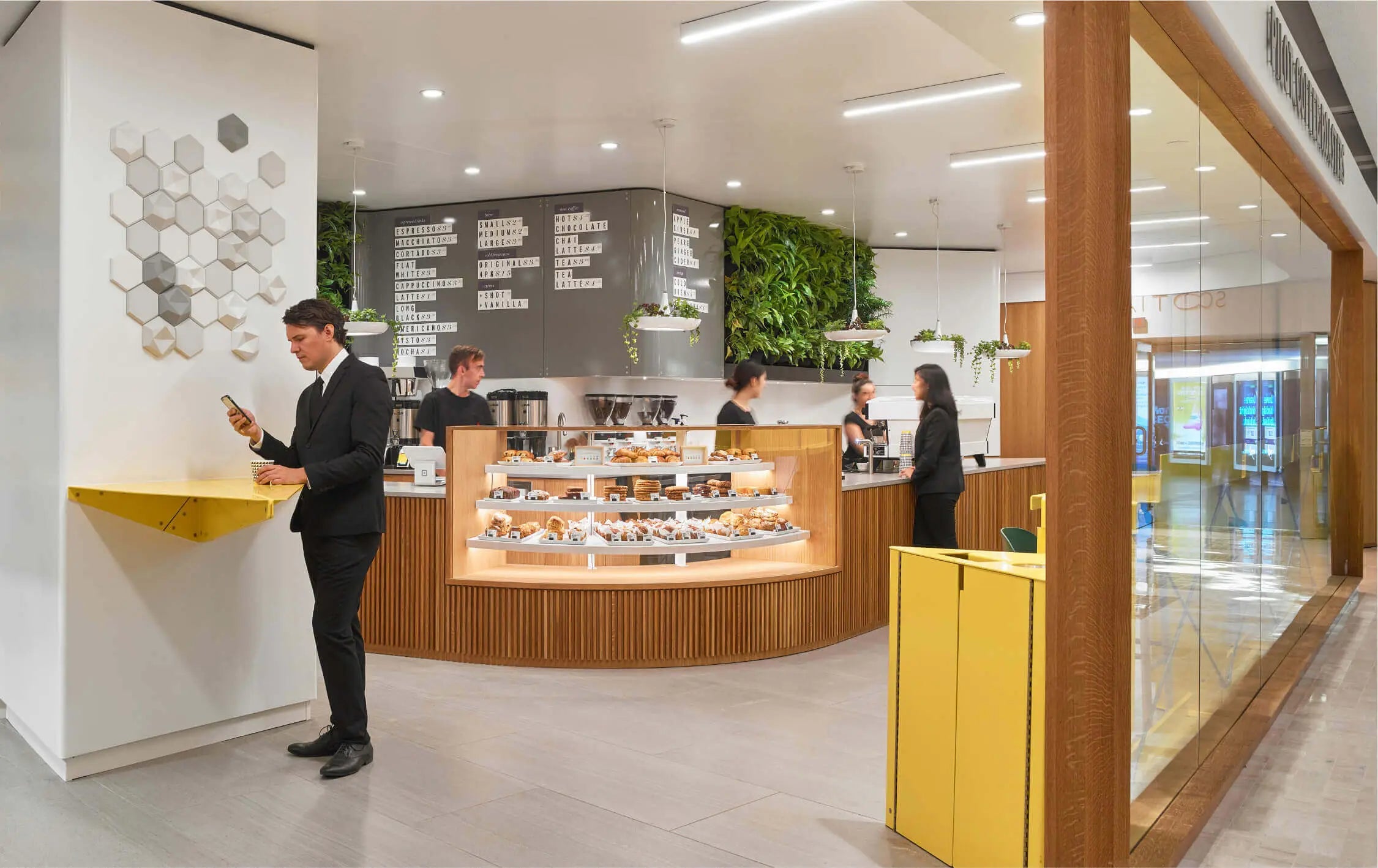 Three baristas can be seen behind the curved wooden bar with one customer placing an order and another drinking a coffee while looking a their phone at a standing table. A live green wall can be seen above the bar beside the grey cafe menu