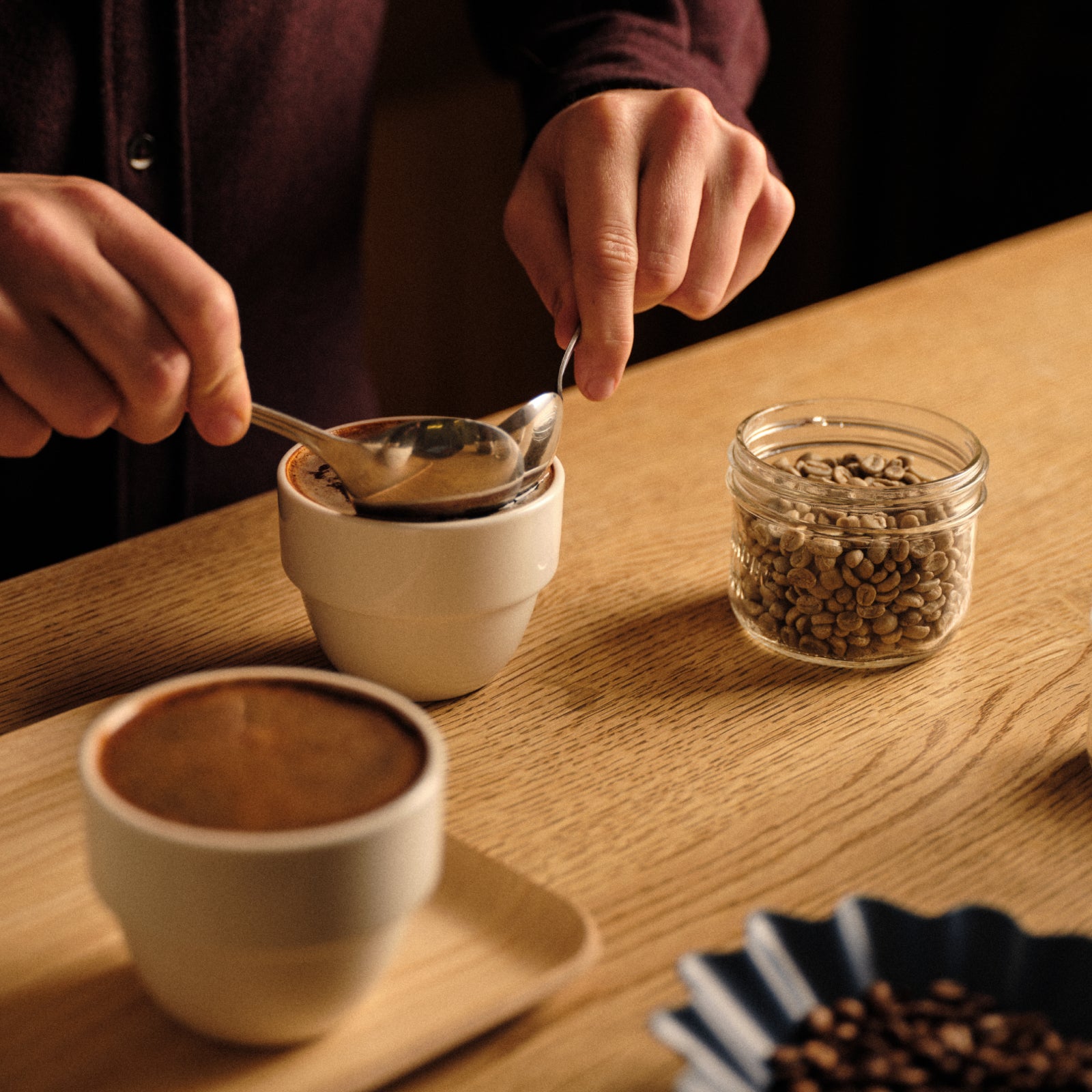 Close up shot of a person's hands breaking the crust during a coffee cupping with another coffee to the left and green beans in an open jar on the right