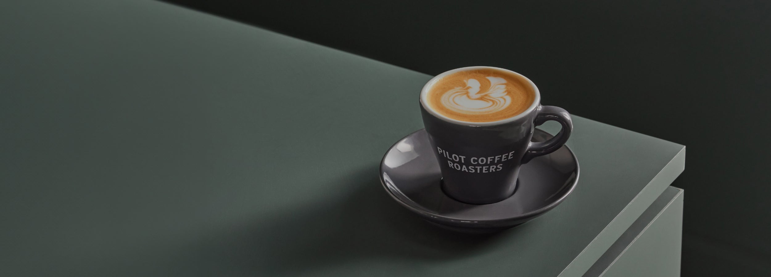 A macchiato in a Pilot Coffee Roasters branded grey cup and saucer sits atop a green countertop. Seahorse latte art on top of the drink.