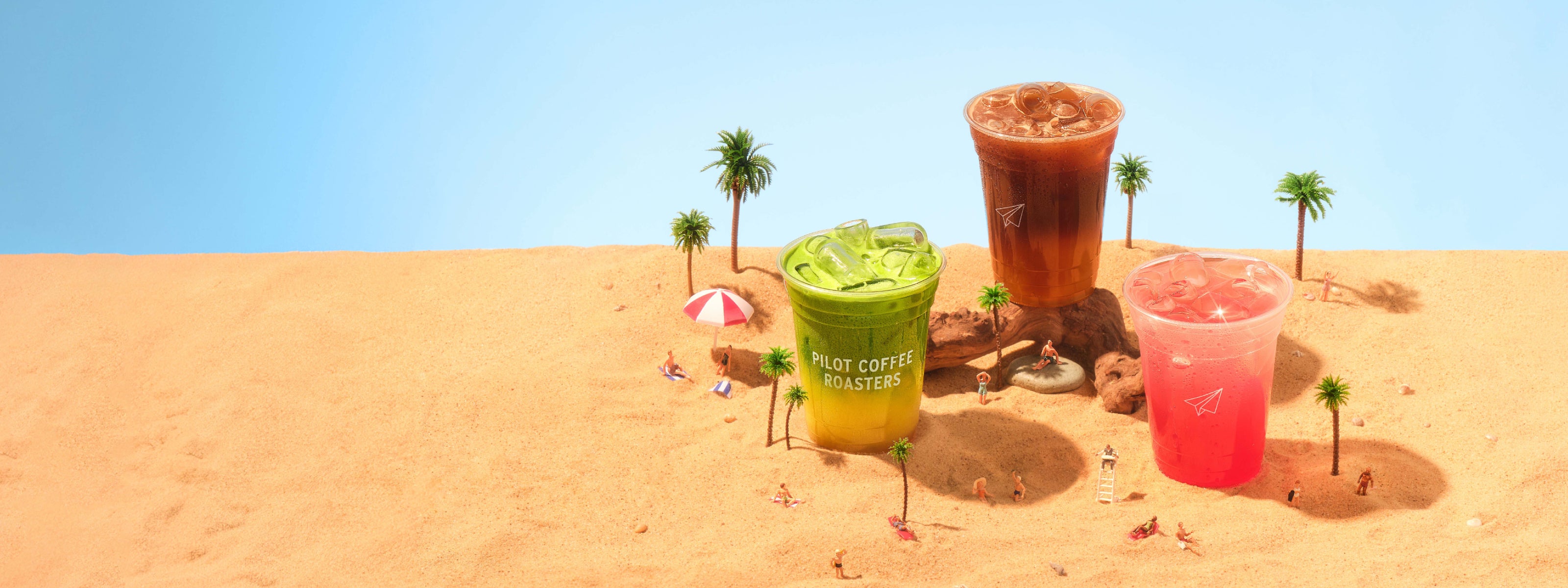 Three summer drinks sit on a diorama of a beach scene with miniatures of trees and beachgoers with a sky blue background. Drink in a transparent to go cup features a gradient of green to yellow with ice on the left. The drink in the center, also iced, is a rich coffee brown and on the right a shining bright pink beverage with ice casts a shadow over the miniatures below. 