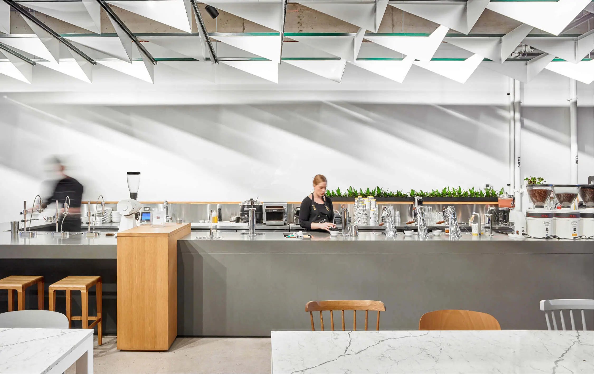 Inside shot of the Ossington Cafe facing the long bar. Two baristas can be seen one in the center steaming milk at the mod bar and another blurred in motion at the pourover station on the left side of the bar. The paper plane architectural feature that covers the ceiling can be seen at the top of the frame.