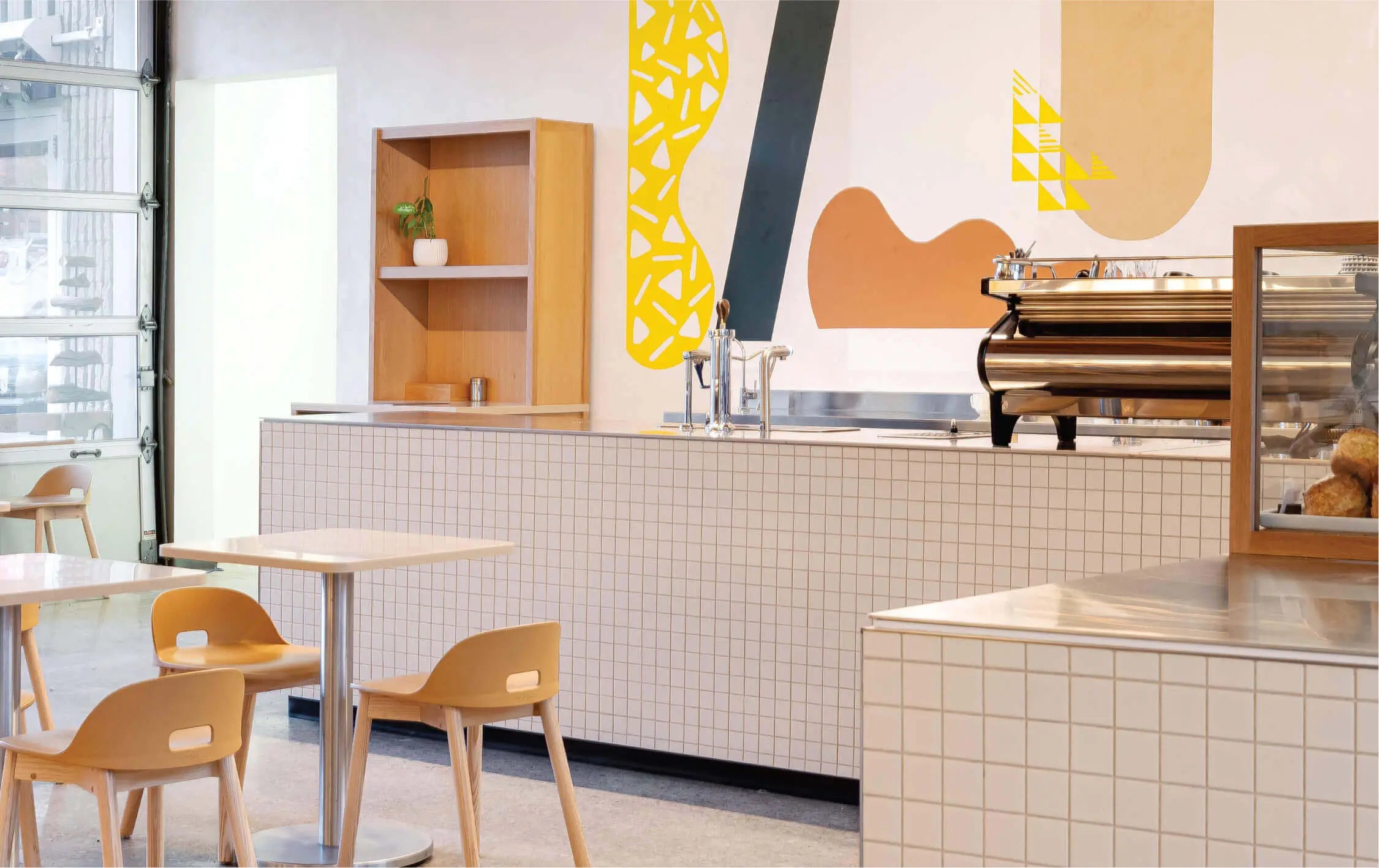 Inside the brightly lit Queen St. East Cafe facing the bar. The Espresso machine, a mod bar steaming station and baked goods display can be seen. Small tables and chairs sit in the main seating area of the cafe