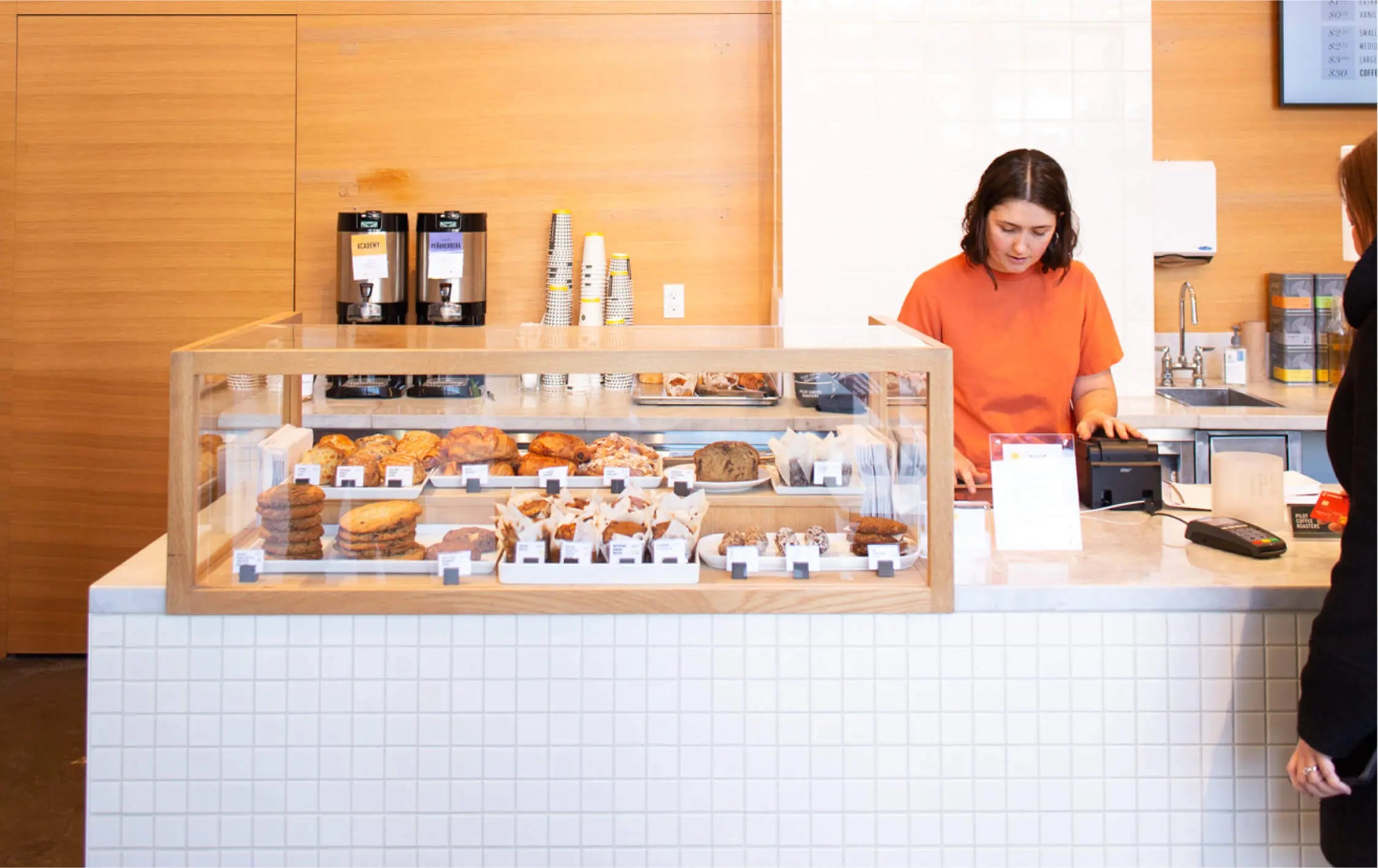 Inside the Richmond St Cafe. A Barista in an orange shirt is checking out a waiting customer. A large glass display of baked goods sits on the bar with brewed coffee and cups behind