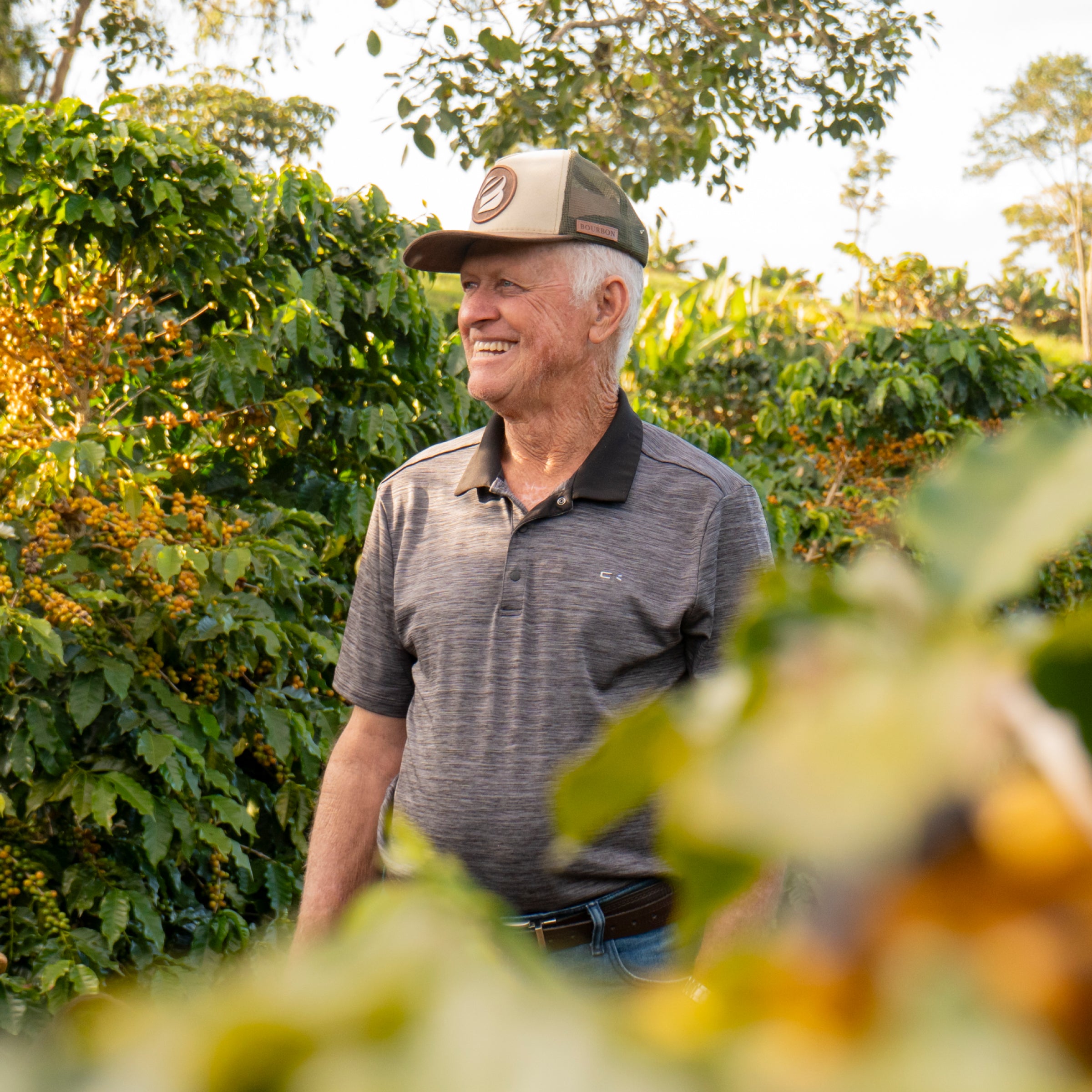 Image shot through blurred coffee plants of Luiz Zanetti smiling surrounded by greenery on his farm. 