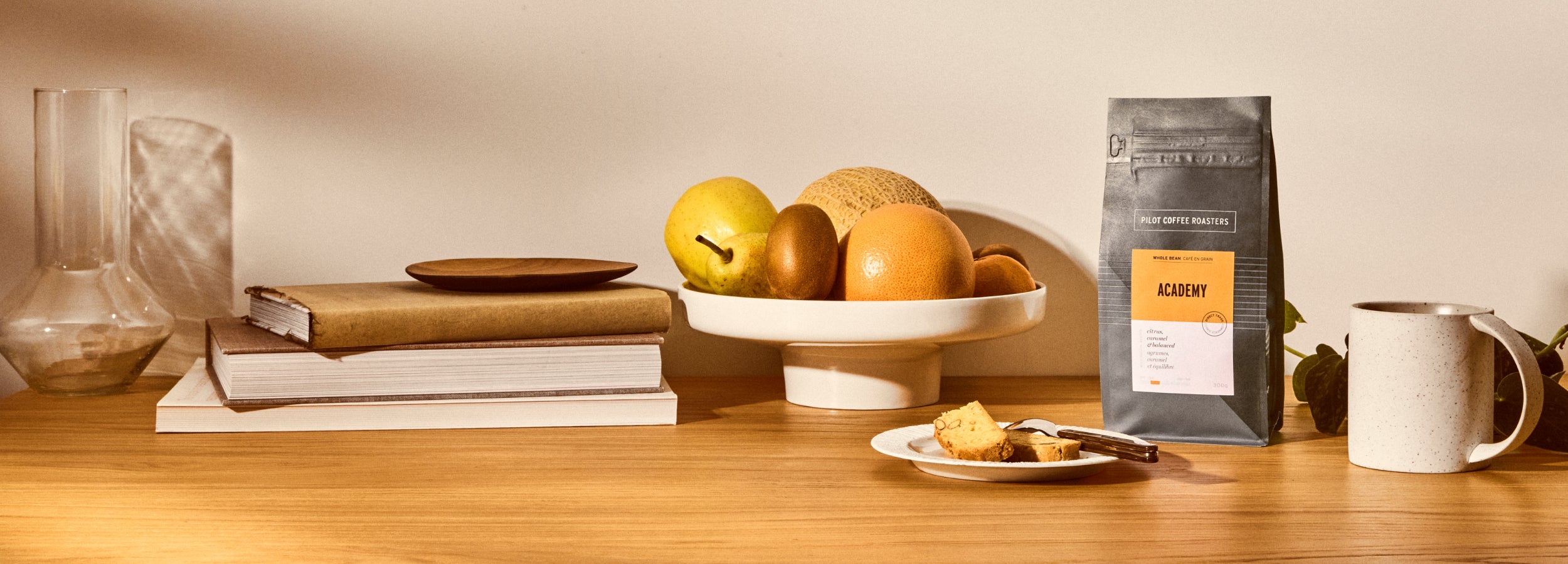 Academy blend on a wood countertop surrounded by a coffee mug to the right, sliced loaf on a saucer with fork in front and cookbooks, a vase, and fruit bowl to the left