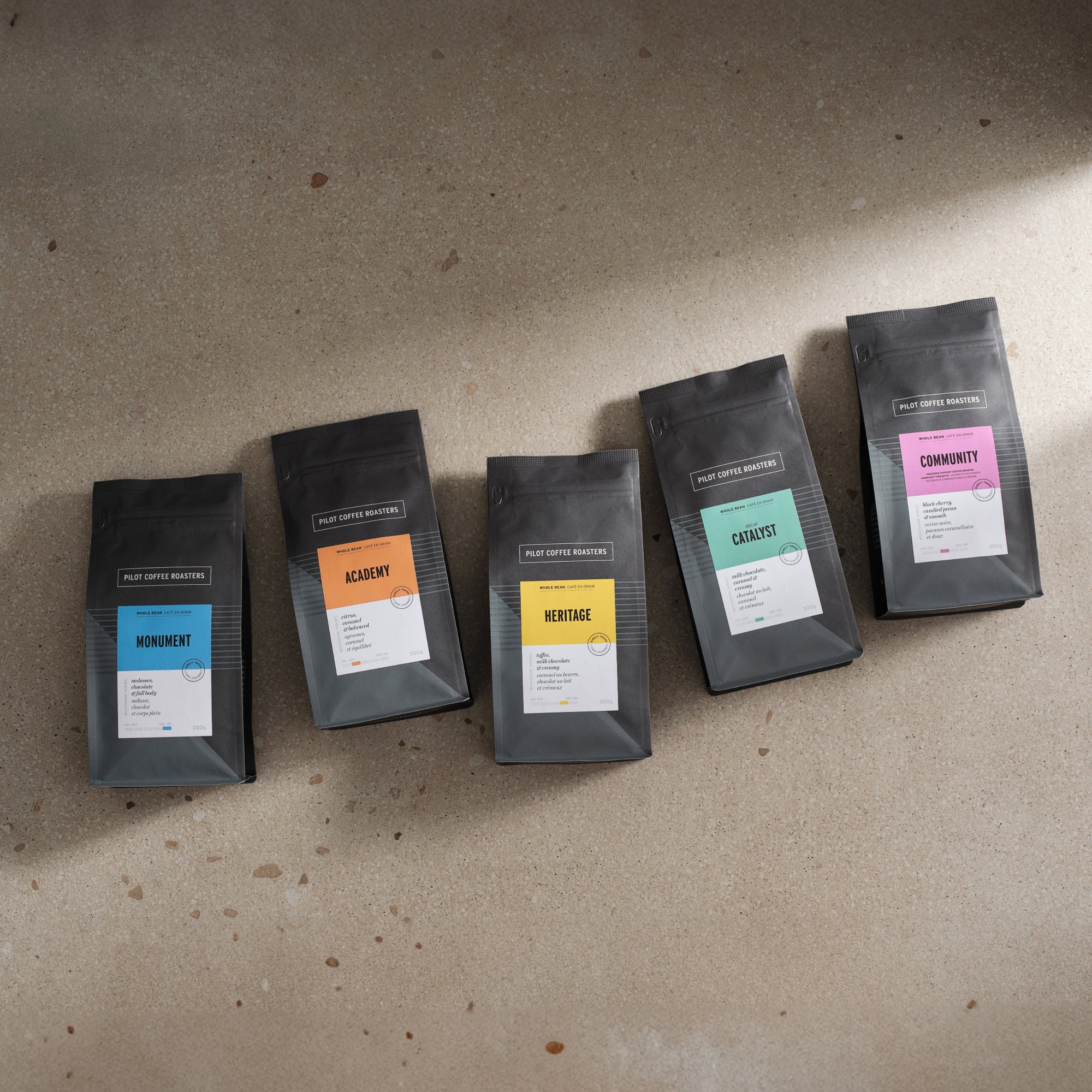 Line up of all five signature Pilot Coffee blends from left to right: Monument blend, Academy Blend, Heritage Blend,  Catalyst Decaf Blend and Community Blend. All laying flat on a shadowy concrete surface