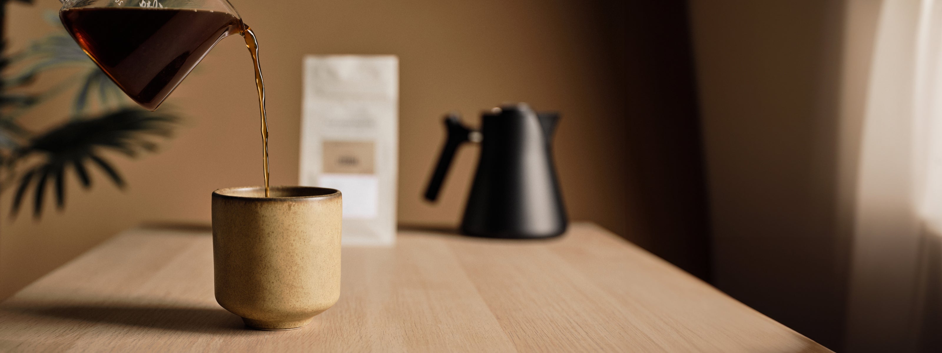 Kalita glass coffee carafe pouring coffee from above into a earth stone coffee cup on a wood counter. A blurred bag of coffee and fellow kettle sit in the background