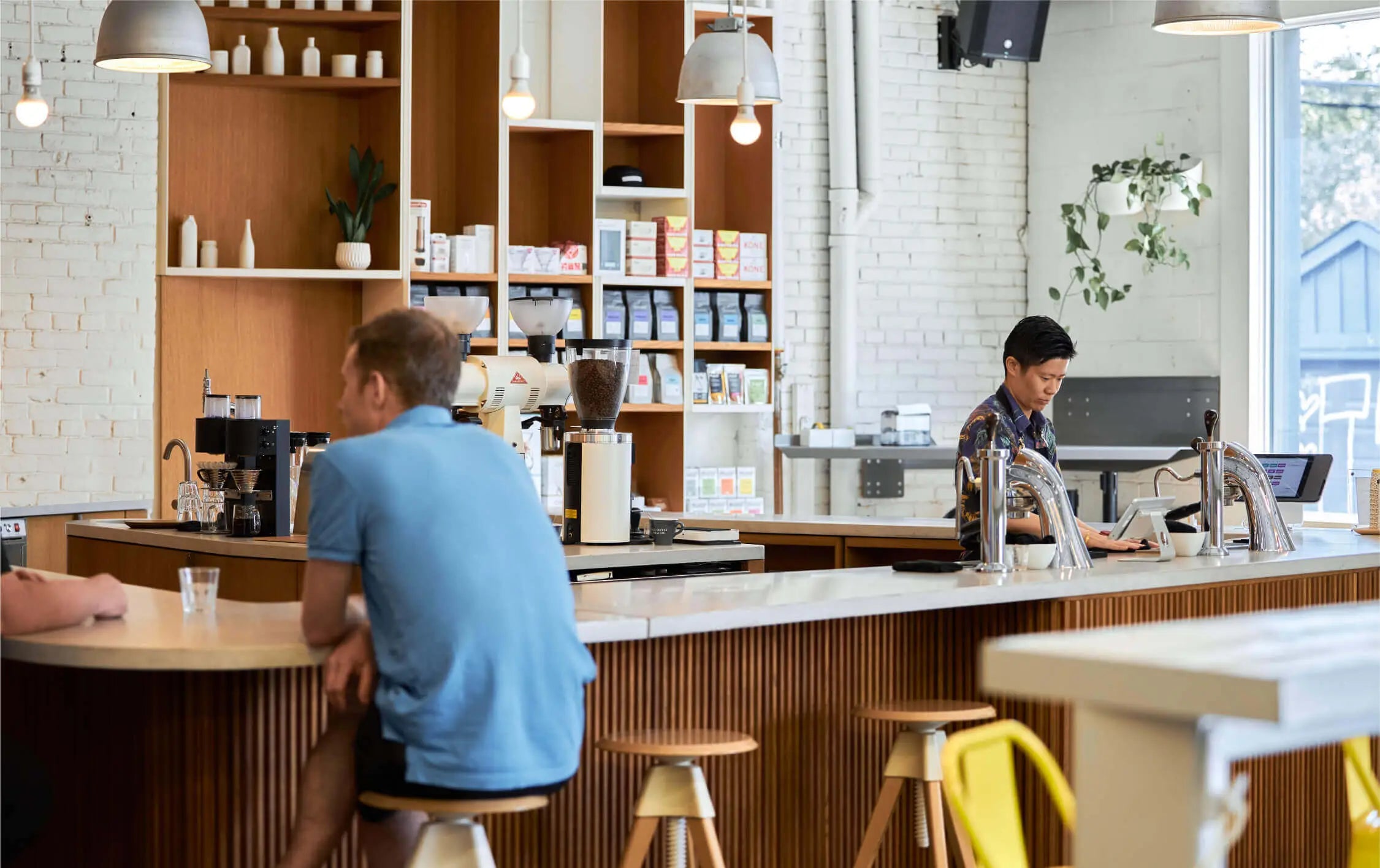 Two customers sit in the brightly lit cafe talking on the left side of the u-shaped wood and concrete bar, one almost completely out of frame. A barista on the right side of the bar is preparing coffee.