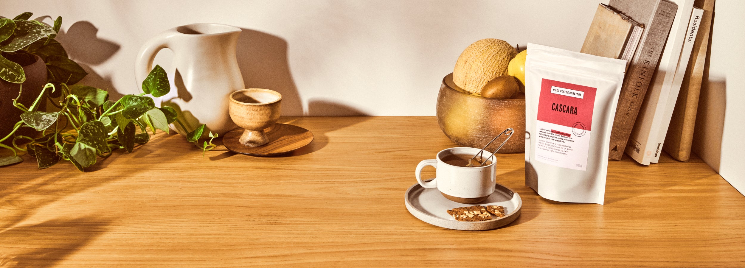 Stylized image of a bag of Cascara tea beside a steeping tea cup and biscup on saucer on a countertop. Vases, plants and cookbooks in the background