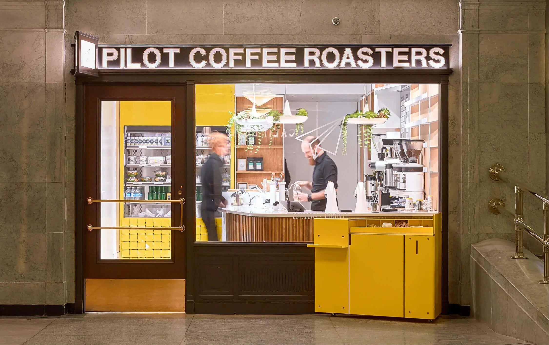 Looking into the glass storefront of the Union Station Cafe. A barista is pouring milk into a takeaway cup while a blurred customer waits in front of the bar.