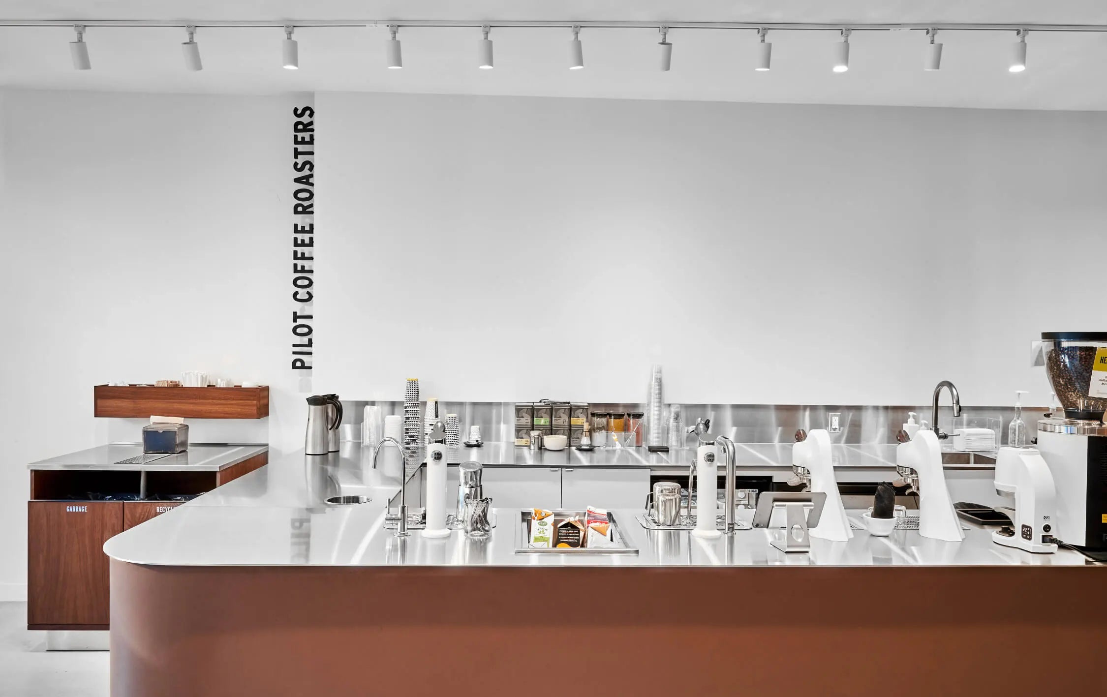 A shot of the curved bar with stainless steel countop and a rich brown front. Two mod bar groupheads and two milk steaming wands are integrated into the worktop. The Pilot Coffee Roasters logo runs vertically up the white wall on the left side of the bar in the background