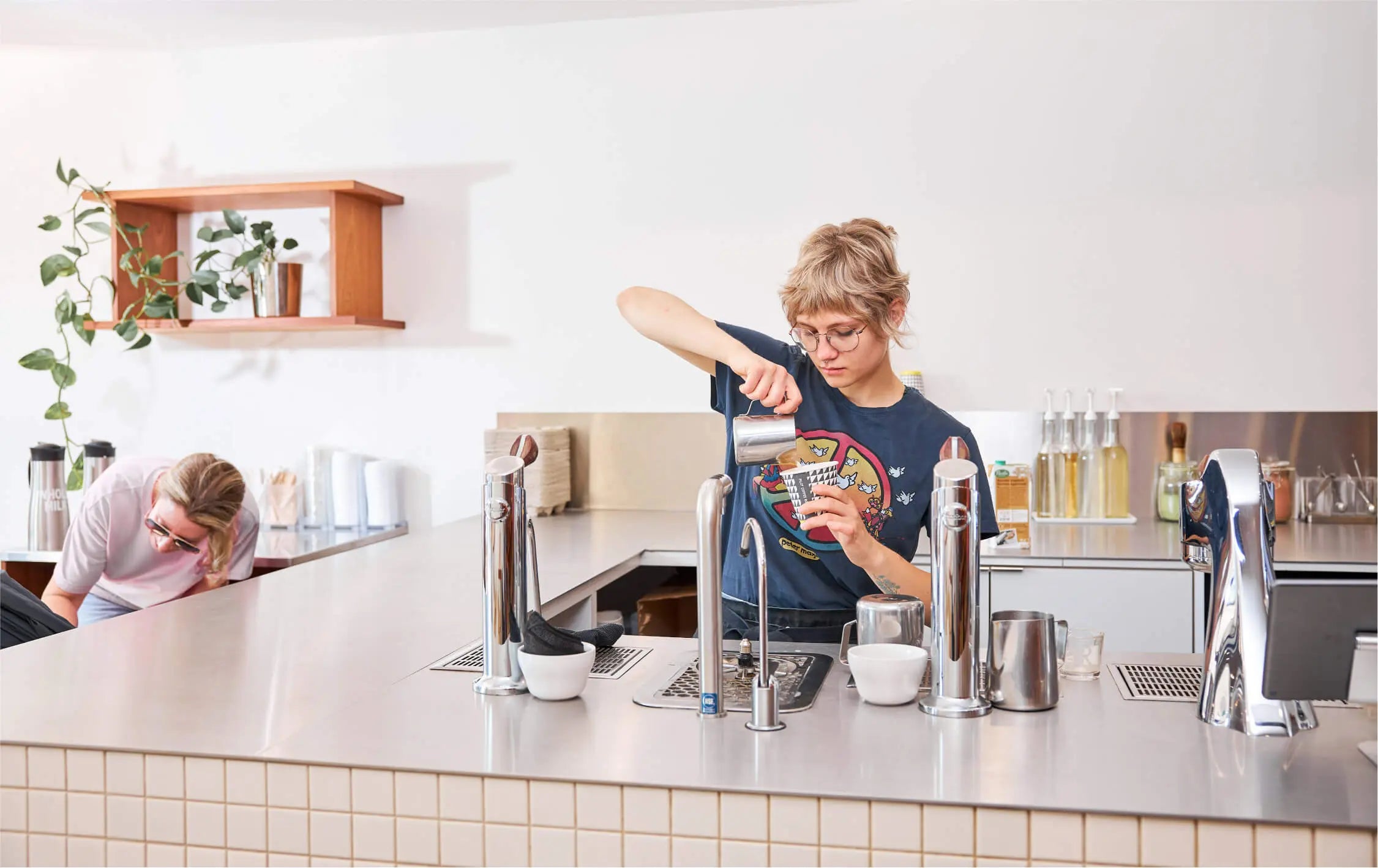 A focused barista pours steamed milk into a latte behind the tile and stainless steel mod bar, while a smiling customer in sunglasses attends to their out of frame child in a stroller. Light is coming in on the left through a window that is out of frame. 